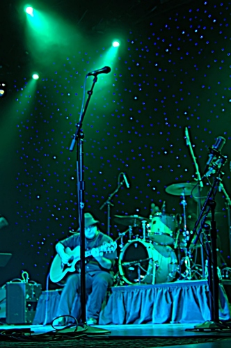 Two Loons For Tea: Guitar Player sitting on the drum riser playing something instrumental.  John Schlick was the Lighting Designer for this show.